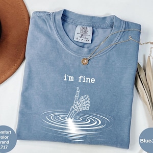 May include: Blue Comfort Colors t-shirt with the text "I'm fine" and a skeleton hand giving a thumbs up. The shirt also has a small tag that says "Comfort Colors Brand 1717". A gold necklace is draped across the shirt.