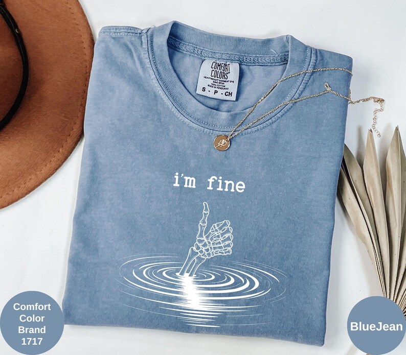 May include: Blue Comfort Colors t-shirt with the text "I'm fine" and a skeleton hand giving a thumbs up. The shirt also has a small tag that says "Comfort Colors Brand 1717". A gold necklace is draped across the shirt.