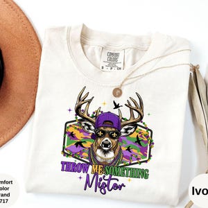 Op de afbeelding: Ivoor kleurig T-shirt met een hert met een zonnebril, een paarse pet en een kralenketting. De afbeelding bevat de tekst "THROW ME SOMETHING Mister" in paars en groen, met een Mardi Gras thema.