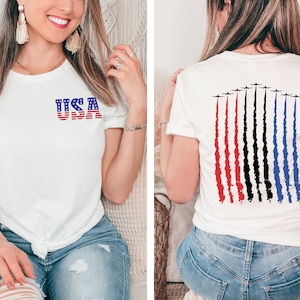 May include: White t-shirt with a red, white, and blue USA graphic on the front and a graphic of red, white, and blue jets flying in formation on the back.