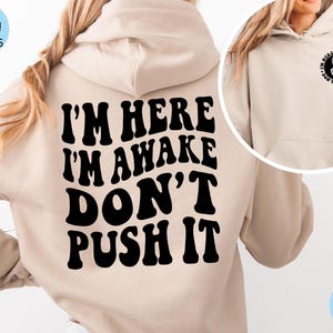 May include: A beige hoodie with black text that reads "I'm here I'm awake don't push it" on the back. The front of the hoodie has a black circle with the text "I'm here I'm awake don't push it" and a peace sign with a skeleton hand inside.