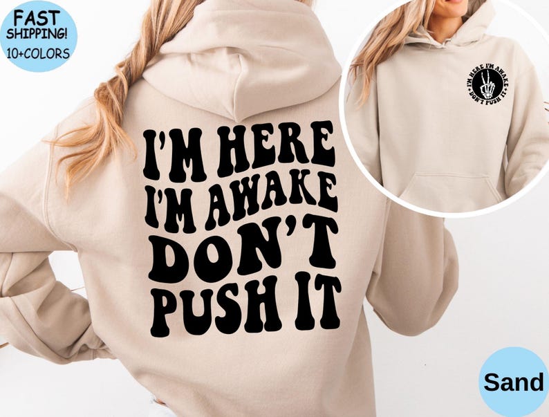 May include: A beige hoodie with black text that reads "I'm here I'm awake don't push it" on the back. The front of the hoodie has a black circle with the text "I'm here I'm awake don't push it" and a peace sign with a skeleton hand inside.