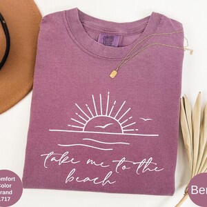 K&ouml;nnte beinhalten: Beerenfarbenes T-Shirt mit wei&szlig;er Grafik von Sonne, Wellen und V&ouml;geln sowie dem Text "Take me to the beach". Eine goldene Halskette liegt auf dem Shirt. Das Shirt ist von der Marke Comfort Colors.