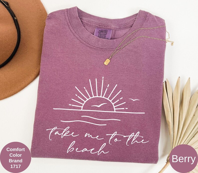 K&ouml;nnte beinhalten: Beerenfarbenes T-Shirt mit wei&szlig;er Grafik von Sonne, Wellen und V&ouml;geln sowie dem Text "Take me to the beach". Eine goldene Halskette liegt auf dem Shirt. Das Shirt ist von der Marke Comfort Colors.