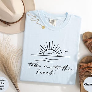 K&ouml;nnte beinhalten: Hellblaues T-Shirt mit schwarzer Grafik eines Sonnenuntergangs &uuml;ber dem Meer und dem Text "Take me to the beach". Das Bild zeigt einen Strohhut, eine goldene Halskette und braune Sandalen.