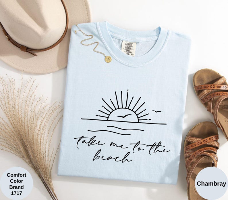 K&ouml;nnte beinhalten: Hellblaues T-Shirt mit schwarzer Grafik eines Sonnenuntergangs &uuml;ber dem Meer und dem Text "Take me to the beach". Das Bild zeigt einen Strohhut, eine goldene Halskette und braune Sandalen.