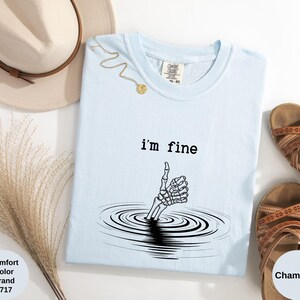 May include: Light blue t-shirt with the text "i'm fine" and a skeleton hand emerging from water, giving a thumbs up. The shirt is a Comfort Colors brand, color Chambray. A gold necklace, a hat, and sandals are also in the image.