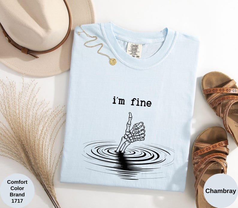 May include: Light blue t-shirt with the text "i'm fine" and a skeleton hand emerging from water, giving a thumbs up. The shirt is a Comfort Colors brand, color Chambray. A gold necklace, a hat, and sandals are also in the image.