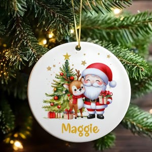 May include: A round white Christmas ornament with a festive design. It features Santa Claus, a reindeer, and a Christmas tree. The ornament has a gold hanging ribbon and the name "Maggie". The design includes red, green, and gold details.