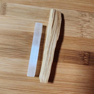 May include: A white selenite wand and a light brown palo santo stick are displayed on a wooden surface. The selenite is rectangular and translucent. The palo santo is a piece of wood with visible grain.