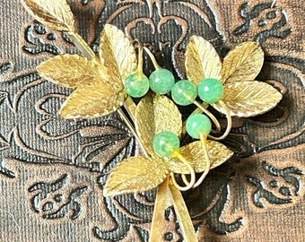May include: A gold-toned brooch with green glass beads and leaf-shaped accents. The brooch is designed to look like a sprig of leaves with berries.