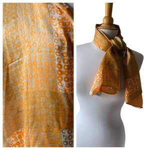May include: A long, orange and white patterned silk scarf with a geometric design. The scarf is tied around a white mannequin's neck.
