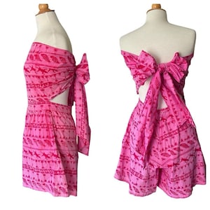 May include: A two-piece pink outfit with a tropical pattern of fish and palm trees. The top is a wrap-around style with a large bow in the back. The shorts have a high waist and a matching print. The outfit is displayed on a mannequin.