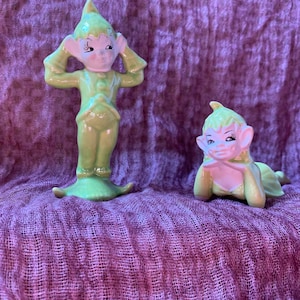 Puede incluir: Dos figuritas de elfo de cerámica verde. Un elfo está de pie con las manos detrás de la cabeza. El otro elfo está acostado con la cabeza apoyada en las manos.