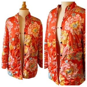 Puede incluir: Chaqueta acolchada con cuello mandarín en un vibrante color naranja, adornada con un gran estampado floral en tonos rosa, amarillo y azul. La chaqueta presenta cierres de botones negros y un brillo sedoso, ofreciendo una estética vintage.