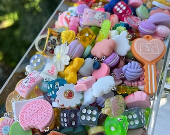 May include: A clear plastic box filled with colorful charms and keychains. The charms include dice, flowers, hearts, clouds, and food items like cookies and macarons. The charms are made of plastic and have a variety of colors, including pink, blue, yellow, green, and purple.
