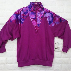 May include: A magenta-colored sweatshirt with a high collar and a V-neck. The upper chest and shoulder area feature a floral pattern in shades of purple and pink. The sweatshirt has long sleeves and a tag that reads "ITANO".