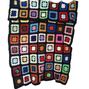 Op de afbeelding: Een kleurrijk gehaakt deken met een zwarte achtergrond en vierkanten in verschillende kleuren, waaronder rood, blauw, groen, geel en bruin.