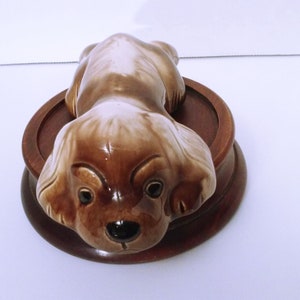 May include: A ceramic figurine of a brown and white dog lying down on a wooden base. The dog has a black nose and brown eyes.