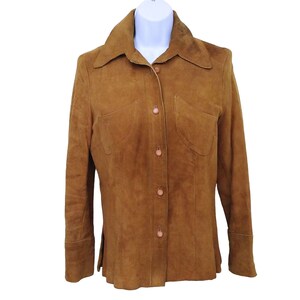 Pode incluir: Casaco de camurça castanho com gola, dois bolsos frontais e botões. O casaco de manga comprida é exibido em um manequim branco. A cor do casaco é um tom quente e terroso.