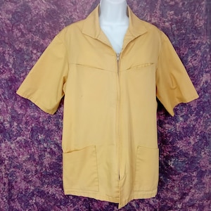 Puede incluir: Chaqueta amarilla clara de manga corta con cierre de cremallera completa. La chaqueta tiene cuello, dos bolsillos delanteros y dos bolsillos en el pecho. La prenda se exhibe en un maniquí blanco sobre un fondo morado y blanco.