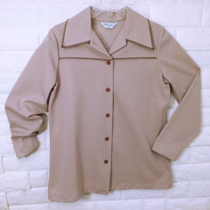 May include: A beige long-sleeved button-up shirt with brown trim around the collar and pockets. The shirt has a pointed collar and six brown buttons down the front.