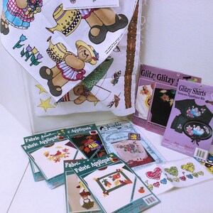 Puede incluir: Surtido de kits de apliques de tela con varios diseños, que incluyen un oso pescando, una niña con flores y un corazón con ángeles. Los kits incluyen instrucciones y apliques termoadhesivos. El texto en el embalaje dice "Fabric Appliqués" y "Glitzy Shirts Iron-on Applique Kit".