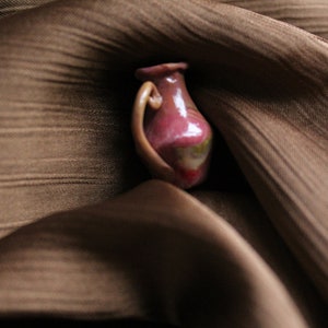 Peut inclure: Un petit vase en céramique avec une anse, présentant un glaçage rose-rouge avec des accents verts et jaunes. Le petit vase est posé sur un fond marron texturé, créant un contraste de couleurs et de textures.