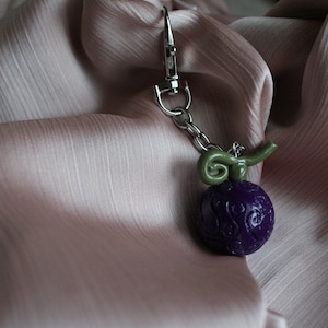 Op de afbeelding: Een paarse, ronde bedel met een groene stengel en een zilverkleurige sluiting. De bedel heeft een gedetailleerd, getextureerd oppervlak en is bevestigd aan een zilveren ketting. De achtergrond is een zachte, roze stof.