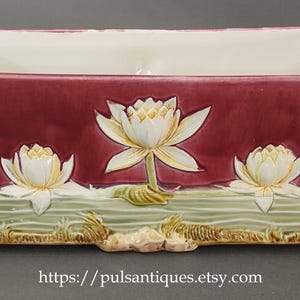 May include: Rectangular ceramic planter with a burgundy exterior and cream interior. The front features three white and yellow water lilies, with green and beige aquatic plants and rocks. The base has a textured design.