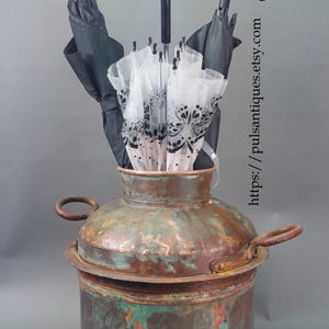 Può includere: Un portombrelli antico in rame con patina consumata, contenente diversi ombrelli. Gli ombrelli includono disegni neri e trasparenti, uno con manico in legno. Il supporto ha due manici.