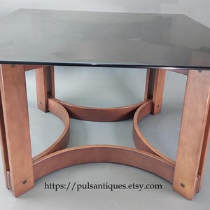 Op de afbeelding: Vierkante eettafel met een donker glazen blad en een houten onderstel. De basis heeft vier gebogen steunen die de poten verbinden. De poten zijn gemaakt van lichtbruin hout. De tafel is ongeveer 91 cm breed en lang.