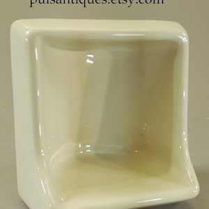 May include: A white ceramic soap dish with a square shape and a slightly curved front edge. The dish has a smooth, glossy finish.
