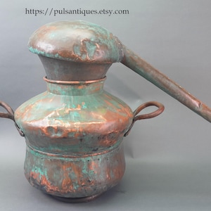 May include: Antique copper still with a rounded base, a bulbous midsection, and a curved spout. The metal has a weathered appearance with green and brown patina, and two handles. The Etsy shop URL is visible at the top.