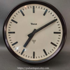 Può includere: Un orologio rotondo vintage con quadrante color crema e lancette e indicatori neri. Il quadrante presenta il marchio "Insa" e un piccolo logo. È racchiuso in una cornice marrone scuro.