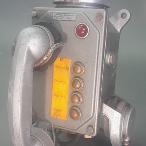 Può includere: Telefono vintage in metallo grigio con campanello, cornetta e pulsanti. Una luce rossa e un'etichetta gialla con testo sono visibili. Il telefono ha un design industriale unico.