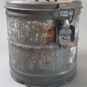 Può includere: Un contenitore in metallo vintage con un coperchio incernierato e un chiavistello. Il contenitore è grigio con la ruggine e ha un'etichetta sbiadita. Il contenitore è alto circa 25 cm.