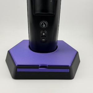 May include: A black electronic device with a smooth, cylindrical handle featuring a circular icon button, a plus sign button, and a minus sign button. The device is seated in a purple and black hexagonal base with a flat, matte finish.