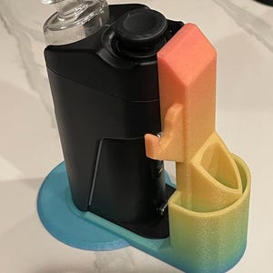 Op de afbeelding: Een zwart elektronisch apparaat met een doorzichtig glazen bovendeel staat in een 3D-geprinte standaard. De standaard heeft een kleurverloop dat van blauw aan de basis overgaat in geel en oranje op de verticale steun en houder.