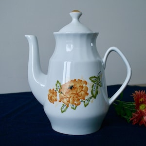 Puede incluir: Tetera de porcelana blanca con un diseño floral con flores naranjas y amarillas y hojas verdes. La tetera tiene una tapa y un asa.