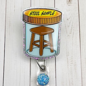 May include: A clear badge reel with a cartoon image of a brown stool inside a jar with the words "STOOL SAMPLE" on top. The reel has a blue glitter accent and a clear plastic clip.