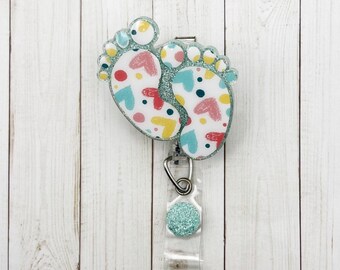 May include: A teal blue baby feet-shaped badge holder with a colorful heart and polka dot pattern. The badge holder has a clear plastic strap and a teal blue glitter button.
