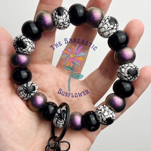 Puede incluir: Un llavero de pulsera con cuentas negras, moradas y blancas. La pulsera tiene un cierre negro y un dije con el texto "The Sarcastic Sunflower". Las cuentas tienen un diseño de telaraña.