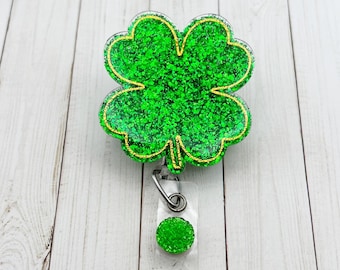 May include: A green glitter four-leaf clover charm with a gold outline attached to a retractable badge holder. The holder has a green glitter circle on the end.