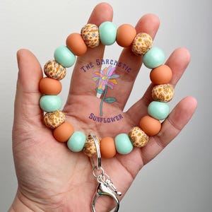 May include: A teal and orange beaded keychain with a floral pattern and a silver clasp. The keychain says "The Sarcastic Sunflower" on it.