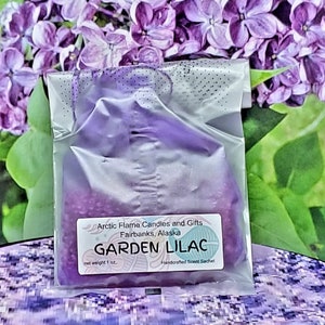 May include: A Garden Lilac scented sachet in a clear plastic bag. The sachet is filled with purple beads and has a label that reads "Arctic Flame Candles and Gifts, Fairbanks, Alaska". The net weight is 1 oz.