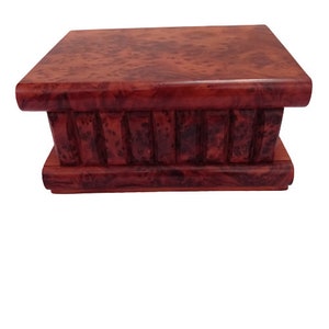 Puede incluir: Una caja de rompecabezas de madera con un acabado marrón oscuro y un patrón de veta de madera decorativo. La caja tiene una tapa con bisagras y una serie de piezas que se entrelazan que deben manipularse para abrirla.
