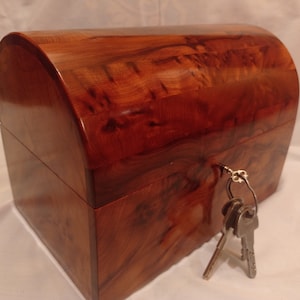 Könnte beinhalten: Eine Holzkiste mit einem gewölbten Deckel, aus edlem rotbraunem Holz mit natürlicher Maserung. Die Kiste hat ein Schlüsselloch und ist mit zwei Schlüsseln gesichert.