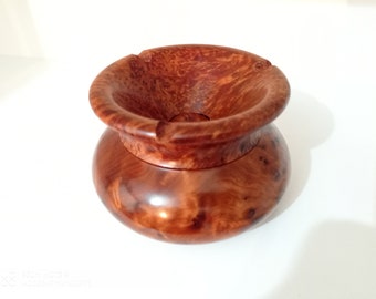 May include: A wooden ashtray with a deep, round bowl and a wide, flared rim. The ashtray is made of a reddish-brown wood with a natural grain pattern.
