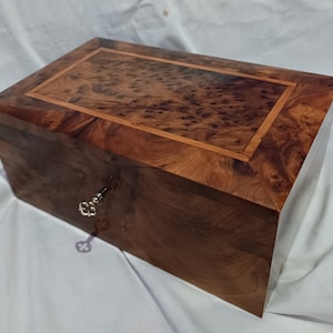 May include: A wooden box with a keyhole and a key in the lock. The box is made of dark wood with a light wood inlay. The box is closed.